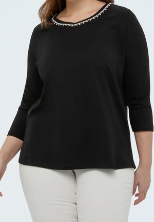 Femme portant un haut noir à manches trois-quarts avec un col rond orné de strass et un pantalon blanc, debout devant un fond uni.