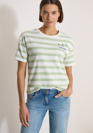 Femme portant un t-shirt rayé vert clair et blanc avec un texte et un jean bleu, se tenant debout avec une main dans la poche sur un fond uni.