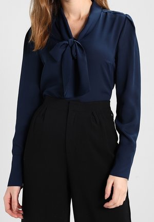 Kvinde iført en mørkeblå bluse med lange ærmer og en knude foran, kombineret med sorte bukser med høj talje.