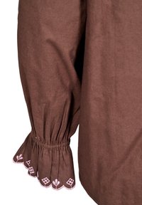 Blusa in tessuto marrone con maniche arricciate. L'orlo delle maniche presenta ricami floreali rosa e un bordo scallop. Texture liscia, vestibilità rilassata.