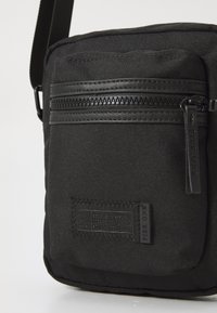 Borsa a tracolla nera con tessuto testurizzato, dotata di una tasca frontale con zip e un'etichetta del marchio. Realizzata con materiali resistenti.
