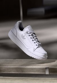 Zapatilla de cuero blanco con puntera redonda, paneles laterales perforados y cordones planos. Presenta un logo sutil y una suela de goma texturizada.