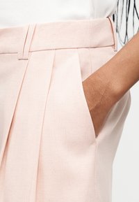 Χέρι φυλαγμένο στην πλαϊνή τσέπη ενός ψηλόμεσου, ανοιχτό ροζ πιετετέ παντελονιού με θηλιές για ζώνη.