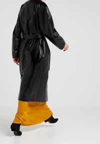 Black, glossy trench coat with long sleeves and a belted waist. Worn over a yellow satin skirt, paired with black high-heeled ankle boots.