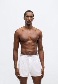 Λευκά βαμβακερά ανδρικά σορτς μποξέρ με ελαστική μέση, που φέρουν το λογότυπο "Calvin Klein" σε μαύρο. Το μοντέλο έχει μυώδη σωματότυπο και δεν φοράει πουκάμισο.