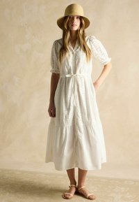 Weißes, gestuftes Midikleid aus Lochstickstoff mit durchgehender Knopfleiste, kurzen Puffärmeln und gebundenem Taillendetail. Getragen mit Sandalen.