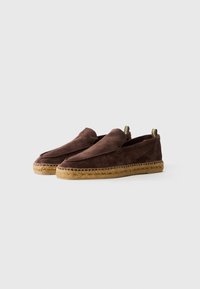 Egy pár barna velúr slip-on espadrille, szőtt juta talppal és szövet fogantyúkkal, fehér háttérrel.