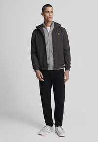 Giacca da uomo, grigia scura, realizzata in materiale morbido con cappuccio, sovrapposta a una felpa leggera grigia con zip. Jeans neri e sneaker bianche.