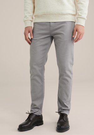 Henkilö, jolla on vaaleanharmaat, kapeat housut ja mustat nyörikengät, seisoo yksinkertaisen neutraalin taustan edessä.