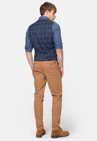 Granatowa kamizelka w kratę o dopasowanym kroju, noszona na niebieskiej koszuli, w połączeniu z beżowymi spodniami slim-fit i brązowymi zamszowymi butami. Subtelne detale z tyłu.