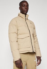 Beige donsjack met hoge kraag, rits aan de voorkant en twee grote zakken. Zachte, gebreide textuur met een minimalistisch ontwerp.