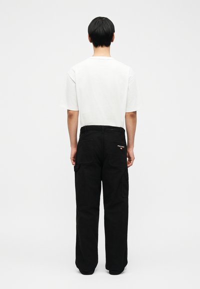 Άνδρας στέκεται με την πλάτη, φοράει λευκό T-shirt και μαύρα παντελόνια cargo με ένα μικρό λογότυπο Polo Sport στην πίσω τσέπη.