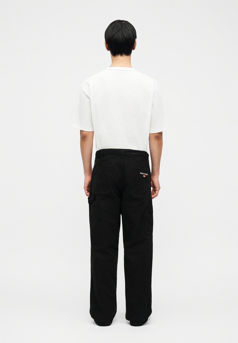 Un homme se tient de dos, portant un T-shirt blanc et un pantalon cargo noir avec un petit logo Polo Sport sur la poche arrière.