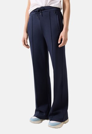 Osoba ubrana w granatowe spodnie z szerokimi nogawkami z przeszyciami z przodu, pas z troczkiem oraz białe i niebieskie trampki, stojąca na prostym tle.