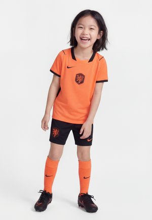 Jeune fille souriante, portant un uniforme de football orange et noir avec un blason de lion, des chaussettes orange et des chaussures à crampons noires, debout sur un fond blanc.