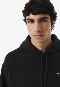 Joven con cabello rizado y oscuro, vistiendo una sudadera negra con cordones y un pequeño texto blanco en el pecho, mirando hacia adelante contra un fondo sencillo.