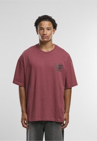 Volné vínové tričko z bavlny s kulatým výstřihem a černou grafickou nášivkou s nápisem „HOME BOY“. Kombinováno se šedými džínami.