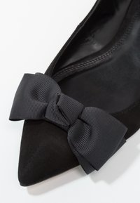 Chaussure plate en suède noir avec un bout pointu et un grand nœud texturé noir à l'avant, dotée d'une doublure intérieure lisse.