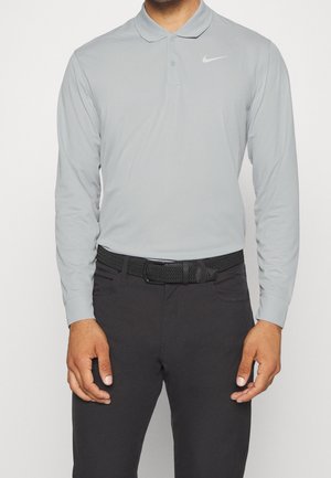 Hombre vistiendo una camisa polo de manga larga color gris claro de Nike con cinturón tejido negro y pantalones oscuros, de pie frente a un fondo liso.