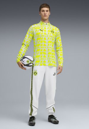 Camicia a maniche lunghe gialla brillante con motivo geometrico, abbinata a pantaloni bianchi con accenti laterali neri e verdi. Incluso un pallone da calcio.