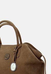 Borsa in suede marrone con doppi manici, targhetta con logo in metallo e medaglione ovale in metallo abbinato appeso a una cinghia di pelle.
