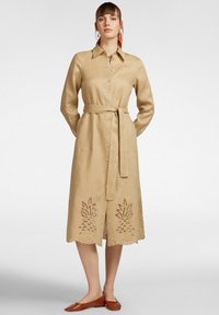 Abito camicia in lino beige con maniche lunghe, chiusura a bottoni, vita con cinturino e design floreale ricamato a intaglio sull'orlo. Scarpe piatte marroni.
