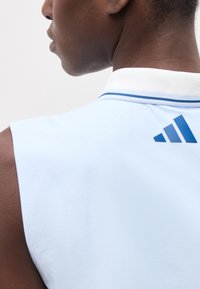 Κοντινό πλάνο ενός ατόμου που φοράει αμάνικο ανοιχτό μπλε πουκάμισο με λευκό γιακά και μπλε λογότυπο Adidas στο πάνω μέρος της πλάτης.