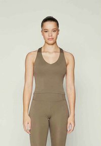 Noor naine, kes kannab ilma varrukateta pruuni sporditopp ja sellele vastavaid leggese, seisab sirgelt heleda neutraalse tausta ees.