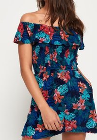 Bloemenprint off-shoulder jumpsuit in donkerblauw met tropische patronen in rood, turquoise en groen. Heeft een geruite halslijn en zakken.