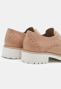 Une paire de chaussures brogue en cuir brun clair avec des perforations décoratives et des semelles en caoutchouc blanc épaisses sur un fond blanc.