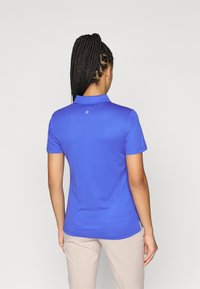 Zila mitruma izsūcoša polo krekls ar tekstūrētu rakstu, īsām piedurknēm un apkakli. Aiz muguras ir neliels logo.