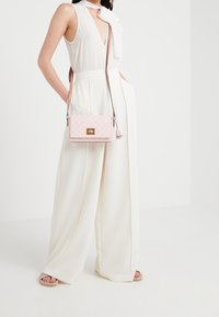 Borsa a tracolla rosa chiaro in materiale testurizzato, con chiusura in metallo dorato e dettaglio a nappina. Indossata con pantaloni bianchi a gamba larga e sandali.