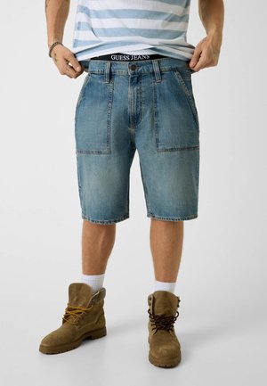 Mann trägt hellblaue Jeansshorts, weiße Socken, braune Schnürstiefel und ein blau-weiß gestreiftes Hemd, hebt den Saum seines Hemdes an.