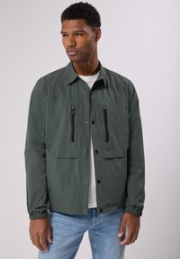 Zelená lehká bunda s dvěma p ředními zipovými kapsami, patentkami, elastickými manžetami a klasickým límcem, nošená přes bílou košili.