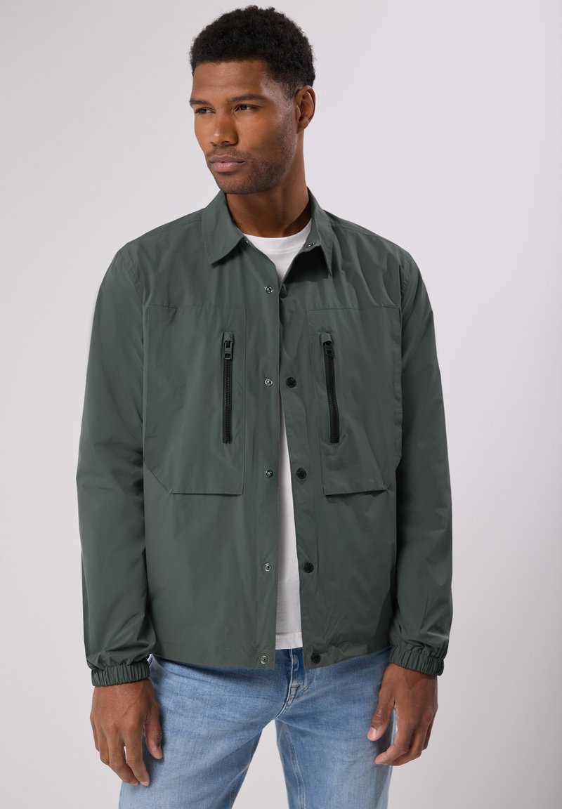 Zelená lehká bunda s dvěma předními zipovými kapsami, patentkami, elastickými manžetami a klasickým límcem, nošená přes bílou košili.