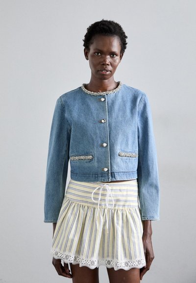 Denim-Jeansjacke im cropped Stil mit Rundhalsausschnitt, silbernen Akzenten und zwei vorderen Taschen. Über einem gelb gestreiften Rock mit Spitzenbesatz getragen.