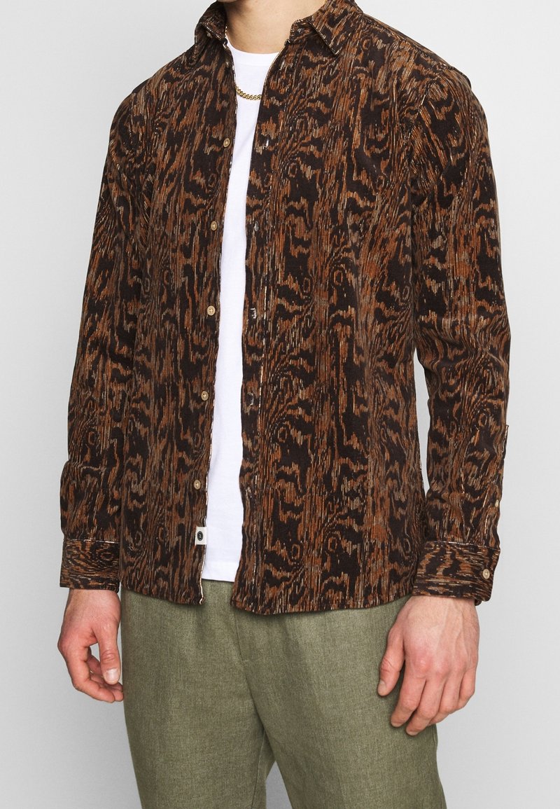 Homme portant une chemise à motifs marron et noir boutonnée sur une chemise blanche avec un pantalon vert olive et un collier chaîne en or.