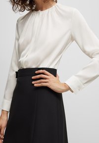Biała bluzka z długim rękawem i marszczonym dekoltem, zestawiona z czarną spódnicą o wysokim stanue. Bluzka posiada delikatne plisy i detale przy mankietach.