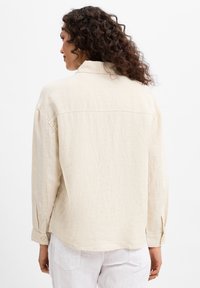 Light beige shirt made of textured fabric, featuring a collared neck, long sleeves, and a slightly oversized fit with a curved hem.
