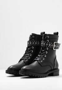 Bottes noires en cuir montant à la cheville avec un design à lacets, une lanière ceinture cloutée, des accents métalliques et une semelle en caoutchouc texturée pour plus de durabilité.