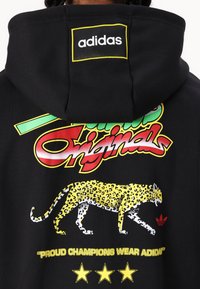 Moleton preto com um gráfico de leopardo amarelo, texto colorido "Os Orgulhosos Campeões Usam adidas" e um pequeno logótipo da adidas na capuz.
