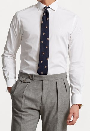 Mand iført hvid skjorte, marineblåt slips med små skjoldmønstre og grå højtaljede plisserede bukser med sidejusteringer.