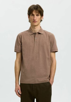 Mężczyzna o brązowych włosach, ubrany w brązową koszulkę polo z krótkim rękawem i ciemnobrązowe spodnie, stojący na jednolitym jasnym tle.