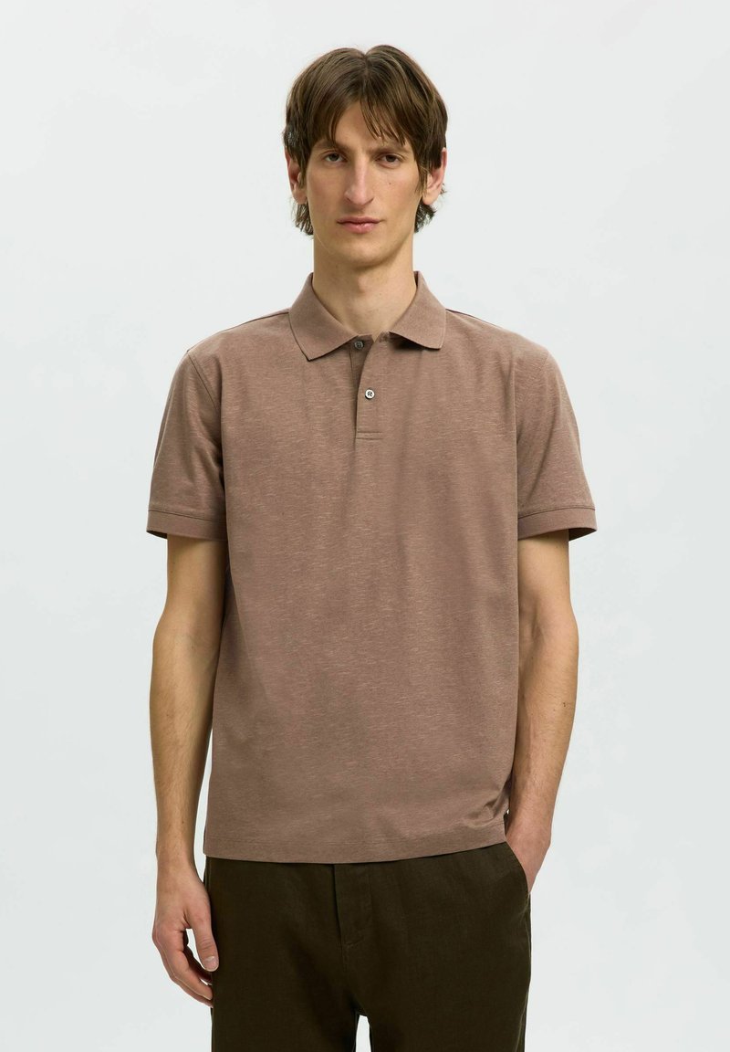 Homme aux cheveux bruns portant un polo marron à manches courtes et un pantalon marron foncé, debout devant un fond clair uni.