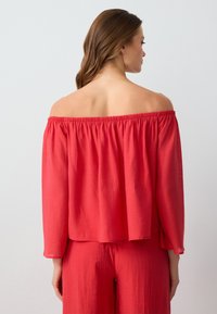 Rotes Off-Shoulder-Oberteil mit gerafftem Ausschnitt, aus leichtem Stoff gefertigt. Lockerer Schnitt mit langen Ärmeln und geradem Saum.