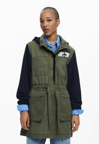 Grüne Canvas-Jacke mit marineblauen Ärmeln, verstellbarem Bund und mehreren Taschen. Mit einem grafischen Design von Händen auf der Brust.