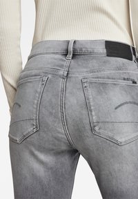 Jeans de mezclilla gris con un corte slim, dos bolsillos traseros con costura curva y una cinturilla texturizada con un parche de logo de cuero negro.
