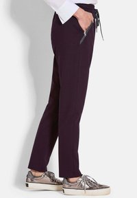 Dunkelviolette, knöchellange Hose mit Kordelzugbund und seitlichen Reißverschlusstaschen, kombiniert mit metallic-silbernen Turnschuhen mit strukturiertem Design.