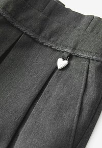 Pantaloni neri plissettati con un tessuto texturizzato. Presentano un piccolo ciondolo a forma di cuore argentato e cuciture visibili sulla vita.