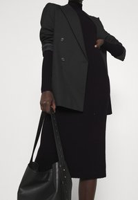 Modella che indossa un abito in maglia a coste nera e un blazer nero, mentre tiene una grande borsa in pelle nera su uno sfondo semplice.